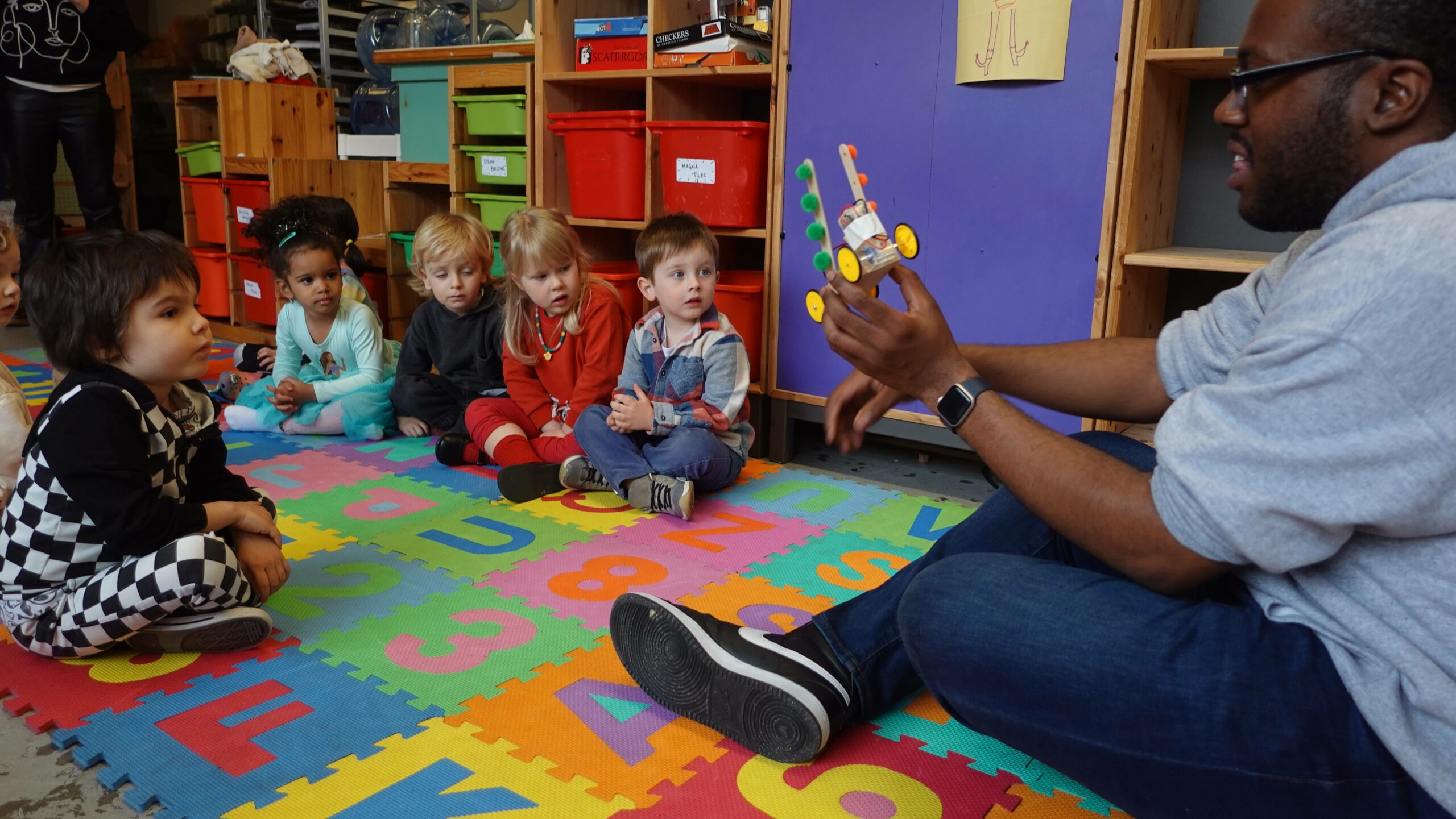 Photo: Students seated on floor looking at teacher, also sitting on floor, holding up a robot.