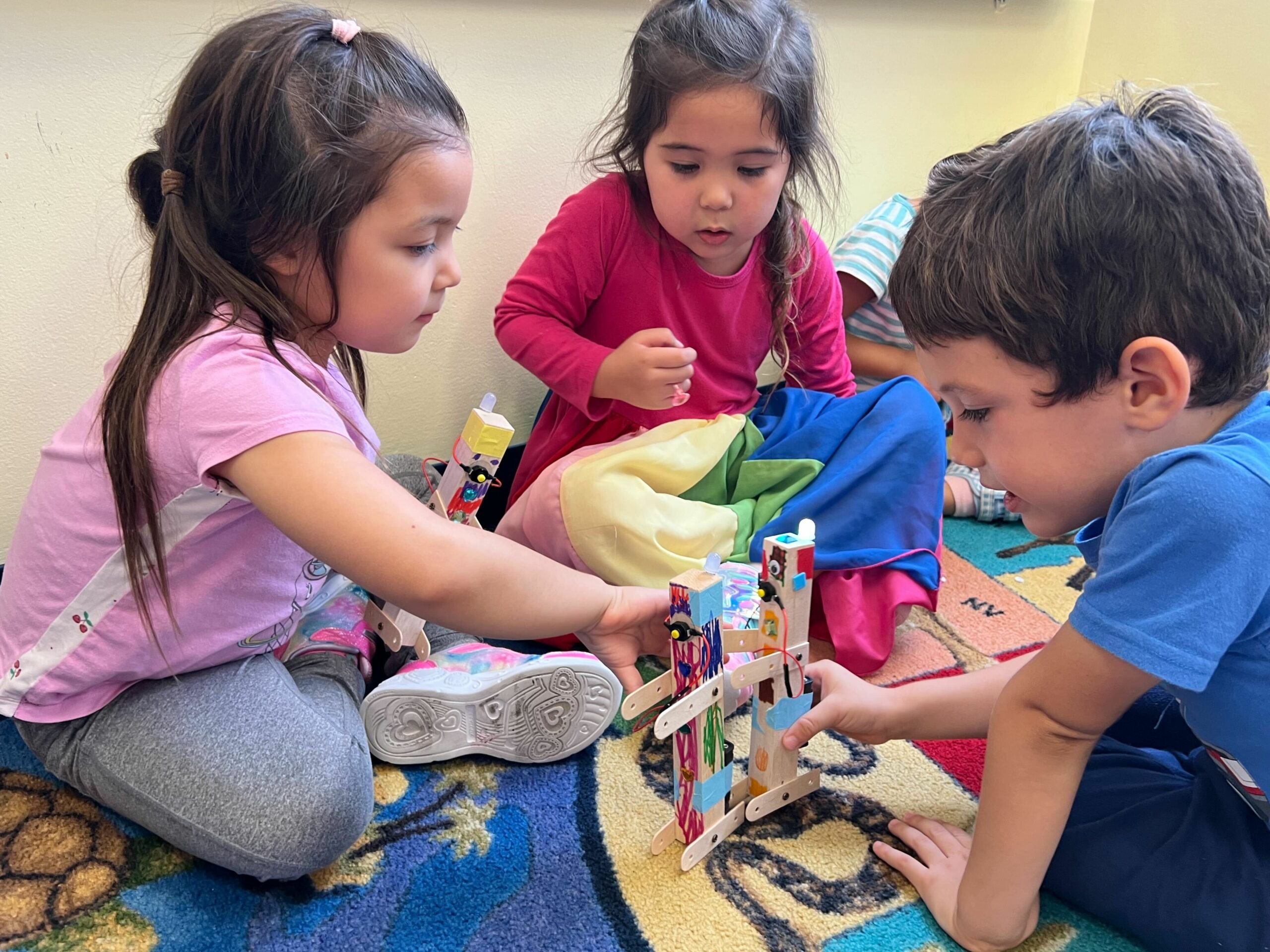 Three kids sit on the floor and play with the robots they just built.