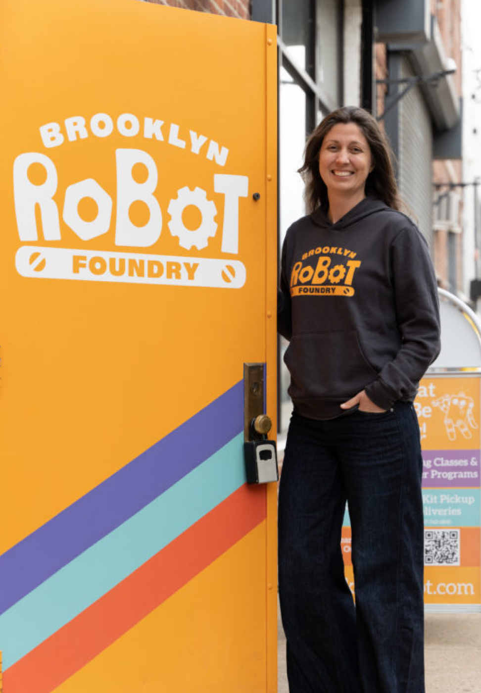 Profile picture (full body) of Jenny Young. She is wearing a grey Brooklyn Robot Foundry sweatshirt and dark pants, standing outside of the orange Brooklyn Robot Foundry door. The blurred city block is behind.