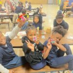 A group of kids sitting at a cafeteria table proudly holding up the little bots that they built.