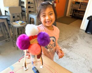 a child holds up the robot they built close to camera. it has 4 large colorful pom poms on it.